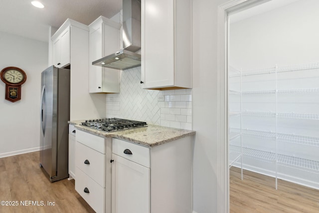 kitchen featuring light wood-type flooring, light stone countertops, stainless steel appliances, white cabinets, and wall chimney exhaust hood