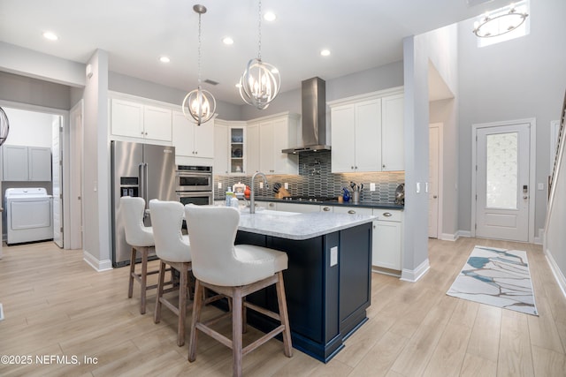 kitchen featuring a center island with sink, appliances with stainless steel finishes, wall chimney range hood, washer / clothes dryer, and white cabinets