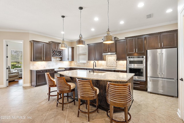 kitchen featuring light stone countertops, hanging light fixtures, stainless steel appliances, a center island with sink, and decorative backsplash