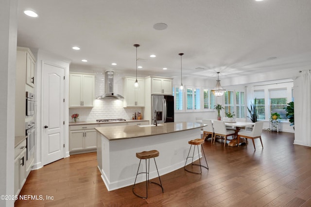 kitchen featuring an island with sink, stainless steel appliances, decorative light fixtures, wall chimney range hood, and white cabinets