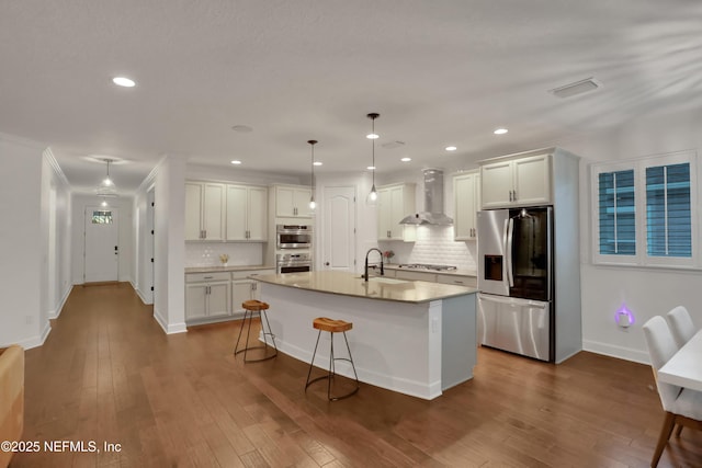 kitchen featuring wall chimney range hood, white cabinetry, hanging light fixtures, a kitchen island with sink, and stainless steel appliances