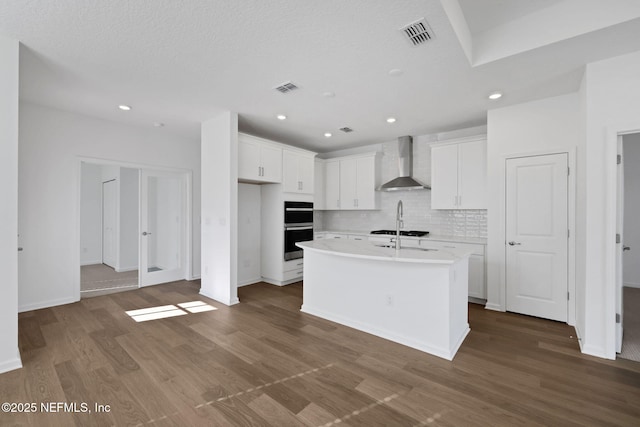 kitchen featuring white cabinetry, wood-type flooring, a center island with sink, double oven, and wall chimney range hood