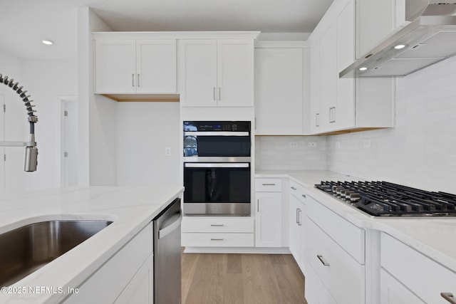 kitchen with white cabinetry, stainless steel appliances, light stone countertops, wall chimney range hood, and light wood-type flooring