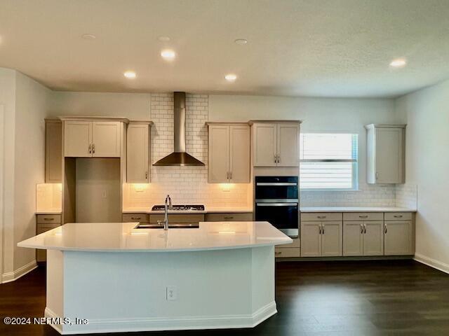 kitchen with dark hardwood / wood-style flooring, double oven, wall chimney range hood, and a kitchen island with sink