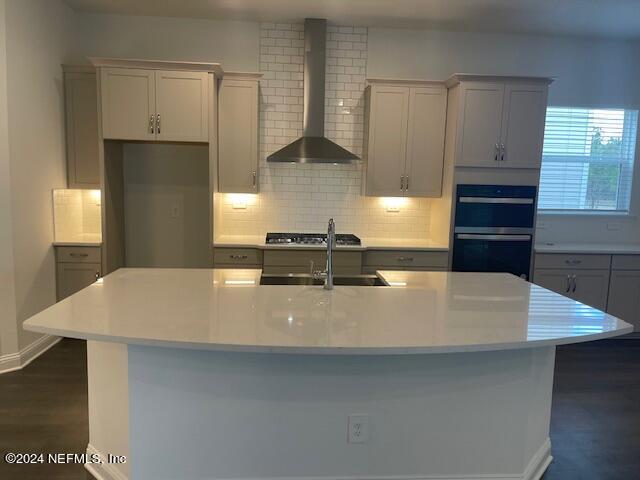 kitchen with gray cabinets, an island with sink, double oven, wall chimney exhaust hood, and sink