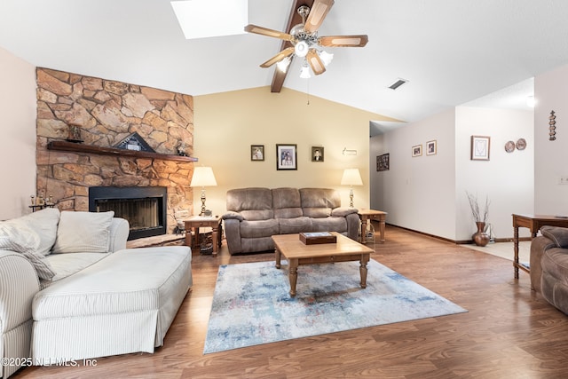 living room featuring ceiling fan, vaulted ceiling with skylight, a stone fireplace, and light wood-type flooring