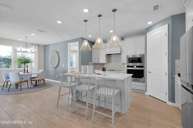 kitchen with a center island with sink, stainless steel appliances, hanging light fixtures, white cabinets, and backsplash