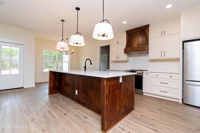 kitchen featuring premium range hood, light hardwood / wood-style floors, a center island with sink, hanging light fixtures, and appliances with stainless steel finishes