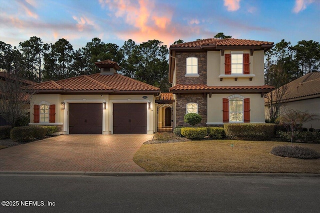 mediterranean / spanish-style house featuring an attached garage, stone siding, a tiled roof, decorative driveway, and stucco siding