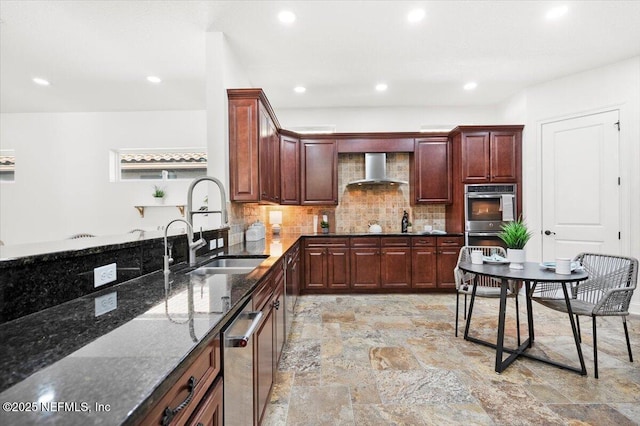 kitchen with appliances with stainless steel finishes, stone finish floor, a sink, wall chimney range hood, and dark stone countertops