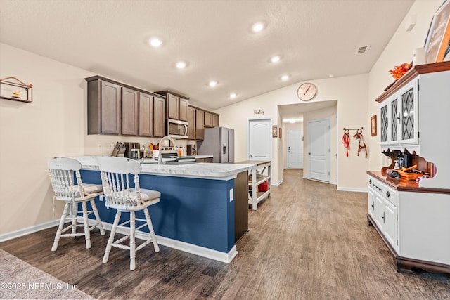 kitchen with kitchen peninsula, hardwood / wood-style floors, appliances with stainless steel finishes, a kitchen breakfast bar, and dark brown cabinets