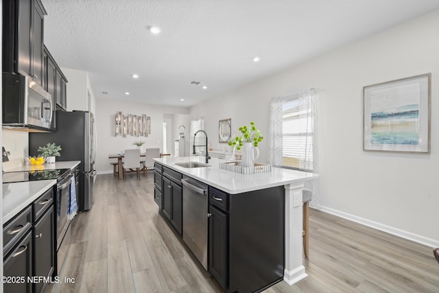 kitchen featuring an island with sink, appliances with stainless steel finishes, light wood-type flooring, a textured ceiling, and sink