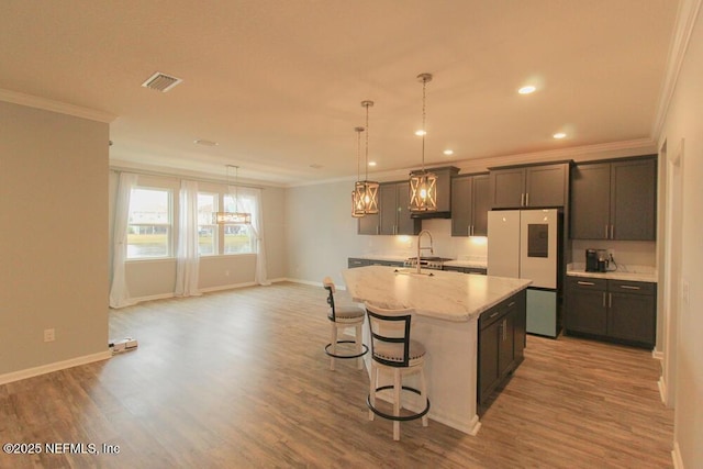 kitchen featuring light stone counters, decorative light fixtures, refrigerator, ornamental molding, and a kitchen island with sink