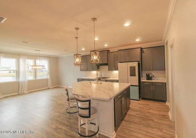 kitchen with light wood-type flooring, stainless steel fridge, pendant lighting, light stone countertops, and a kitchen island with sink