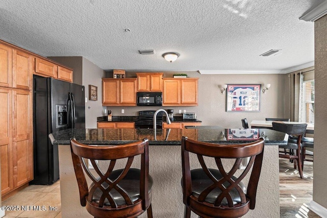 kitchen featuring a center island with sink, black appliances, crown molding, light tile patterned floors, and dark stone counters