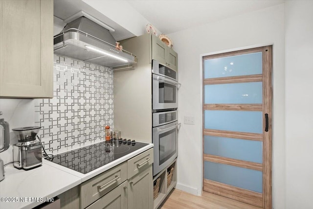 kitchen featuring wall chimney range hood, light hardwood / wood-style flooring, double oven, black electric cooktop, and decorative backsplash