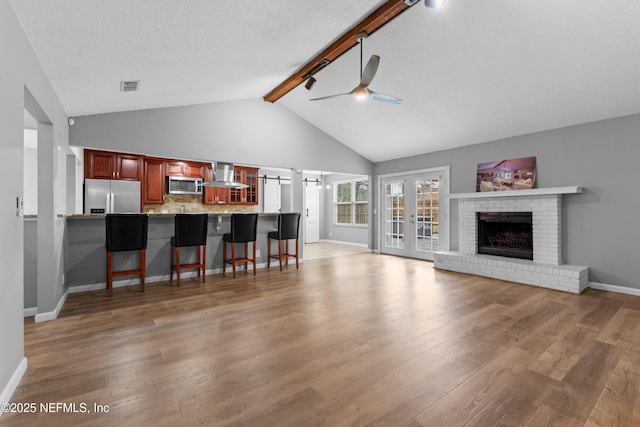 living room with ceiling fan, french doors, light wood-type flooring, lofted ceiling with beams, and a brick fireplace