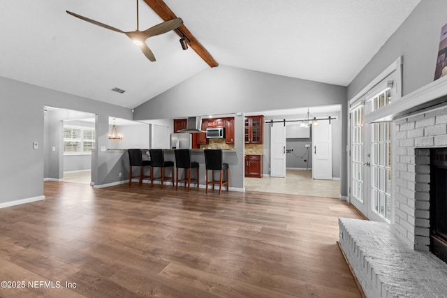 living room featuring ceiling fan with notable chandelier, hardwood / wood-style flooring, a brick fireplace, beam ceiling, and a barn door