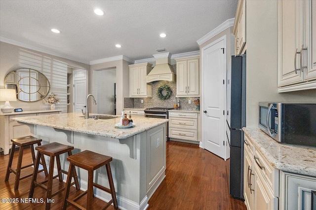 kitchen featuring custom exhaust hood, appliances with stainless steel finishes, an island with sink, ornamental molding, and light stone counters