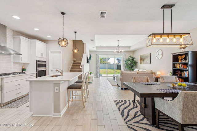 kitchen featuring pendant lighting, wall chimney range hood, a breakfast bar area, a kitchen island with sink, and white cabinetry