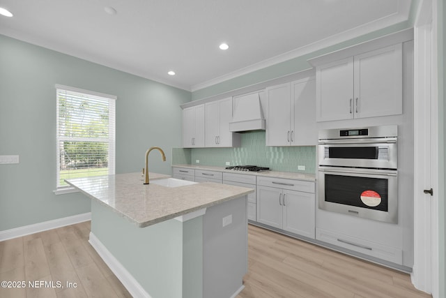 kitchen with custom exhaust hood, an island with sink, sink, and white cabinets