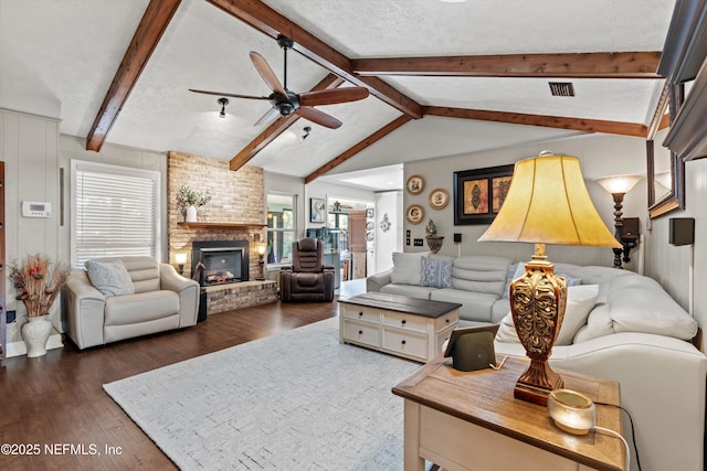 living room with vaulted ceiling with beams, ceiling fan, a brick fireplace, dark wood-type flooring, and a textured ceiling