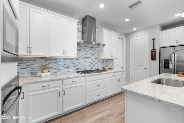 kitchen featuring light stone counters, appliances with stainless steel finishes, white cabinets, wall chimney range hood, and backsplash