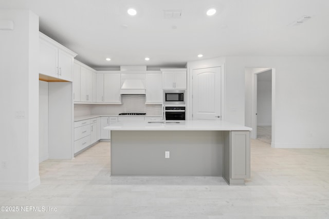 kitchen with black appliances, an island with sink, white cabinets, decorative backsplash, and custom exhaust hood