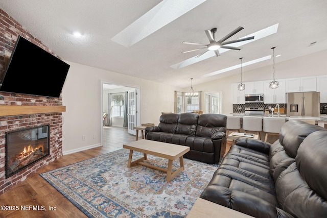 living room with ceiling fan, vaulted ceiling with skylight, a brick fireplace, wood-type flooring, and sink