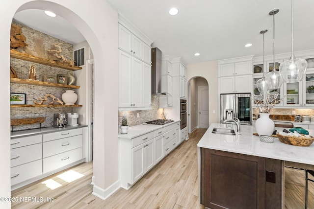 kitchen featuring sink, hanging light fixtures, stainless steel appliances, a center island with sink, and wall chimney range hood