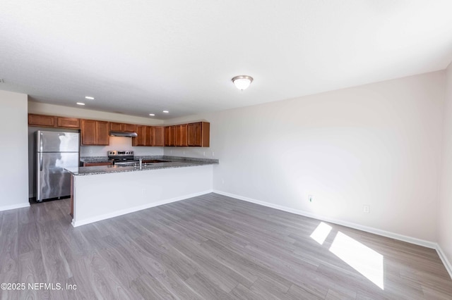 kitchen featuring under cabinet range hood, wood finished floors, stainless steel appliances, a peninsula, and brown cabinetry