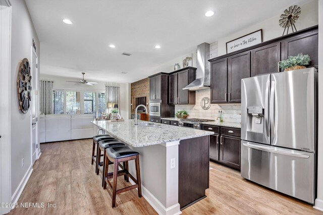 kitchen featuring dark brown cabinetry, stainless steel appliances, a center island with sink, and wall chimney range hood