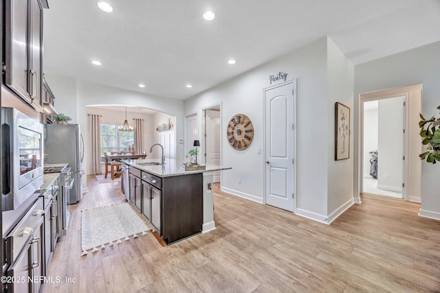 kitchen featuring light stone counters, an island with sink, dark brown cabinets, and light wood-type flooring