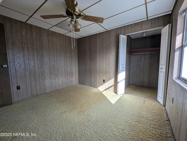 unfurnished bedroom featuring light carpet, wooden walls, a closet, ceiling fan, and a drop ceiling