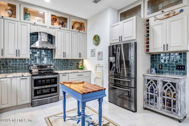 kitchen featuring black fridge, white cabinetry, double oven range, light stone countertops, and wall chimney range hood