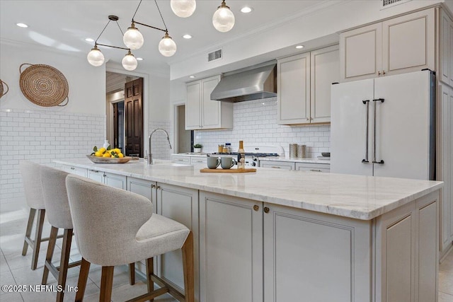 kitchen with light stone countertops, decorative backsplash, a kitchen island with sink, and wall chimney range hood