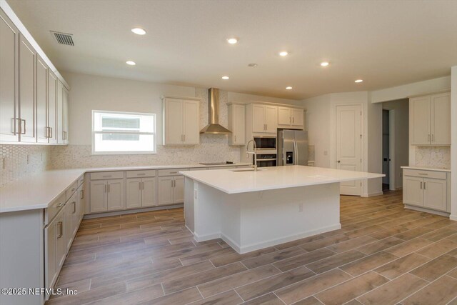 kitchen with appliances with stainless steel finishes, wall chimney exhaust hood, recessed lighting, backsplash, and light wood finished floors