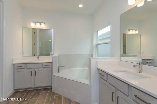 bathroom with a stall shower, a bath, two vanities, wood finished floors, and recessed lighting