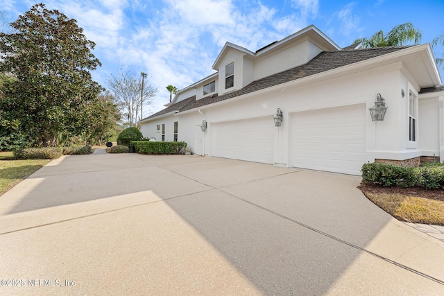 view of side of property with a garage, concrete driveway, a shingled roof, and stucco siding