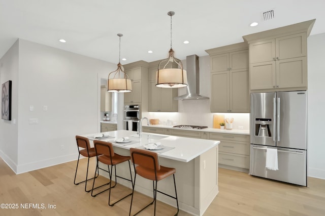 kitchen featuring light hardwood / wood-style floors, appliances with stainless steel finishes, a kitchen island with sink, wall chimney range hood, and sink