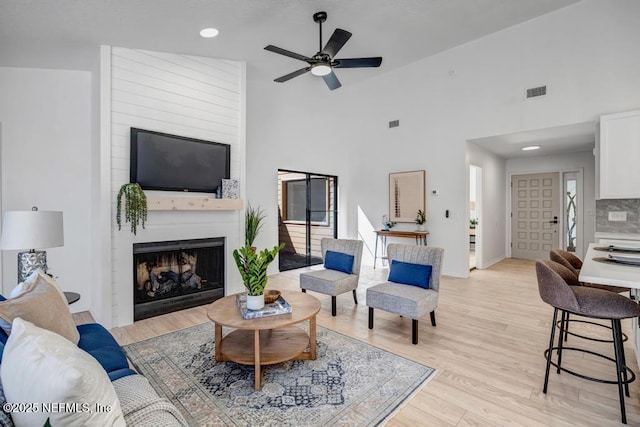 living room with ceiling fan, a large fireplace, high vaulted ceiling, and light wood-type flooring