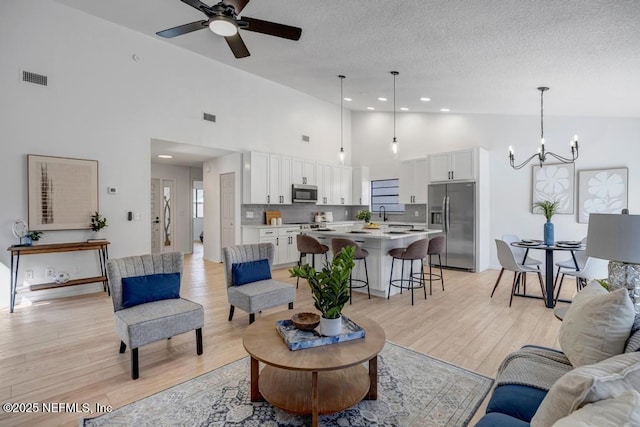 living room with ceiling fan with notable chandelier, high vaulted ceiling, sink, light hardwood / wood-style floors, and a textured ceiling