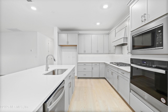 kitchen with sink, gray cabinets, stainless steel appliances, custom range hood, and light wood-type flooring