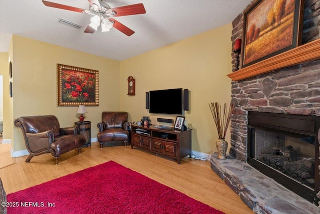 living room featuring ceiling fan, a stone fireplace, a textured ceiling, and light wood-type flooring