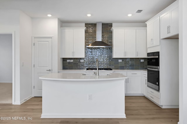 kitchen featuring an island with sink, white cabinets, black double oven, wall chimney range hood, and light wood-type flooring