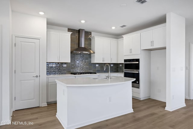 kitchen featuring wall chimney exhaust hood, sink, white cabinetry, a center island with sink, and black appliances