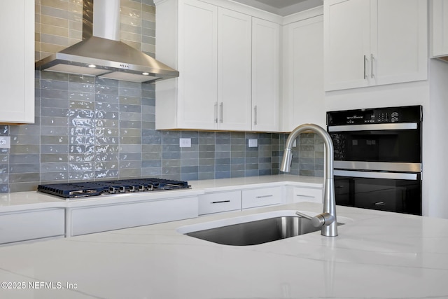 kitchen featuring sink, multiple ovens, light stone counters, white cabinets, and wall chimney exhaust hood