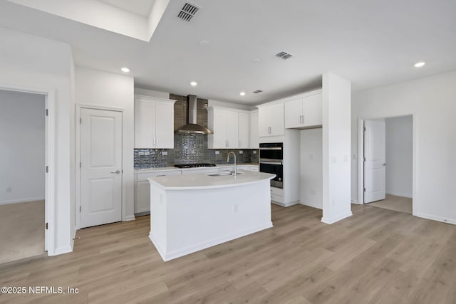 kitchen featuring wall chimney exhaust hood, sink, white cabinetry, a kitchen island with sink, and black appliances