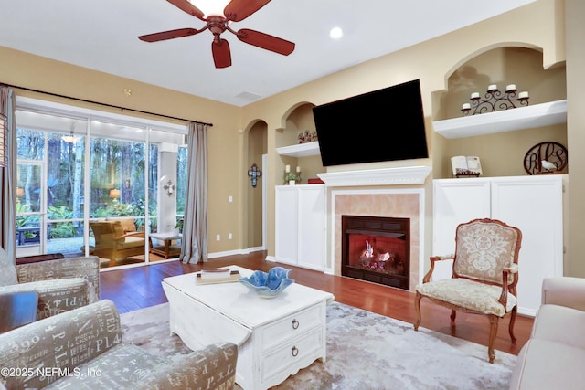 living room with ceiling fan, wood-type flooring, built in features, and a tiled fireplace