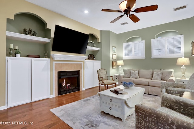 living room featuring ceiling fan, built in shelves, a tile fireplace, and light wood-type flooring